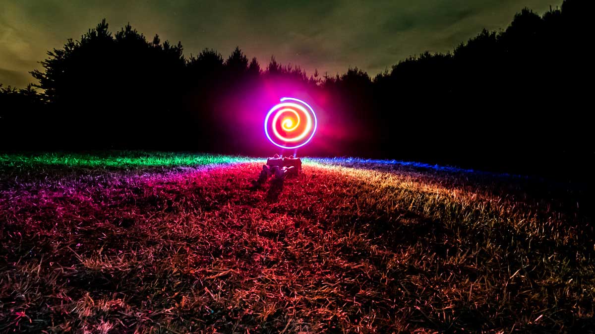 Night photo using Neon lights and long exposure to create a circle image of light paint with two people laying the in grass below the light.